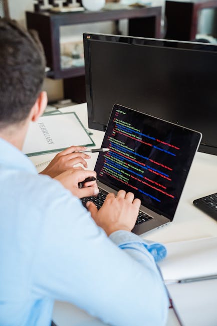 A young man with dark, curly hair is working intently at a desk in a modern office setting, using a keyboard and mouse to operate his computer. The monitor displays lines of code, indicating programmi