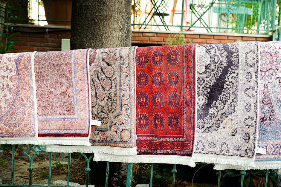 An outdoor urban street scene featuring a building façade with three colorful oriental rugs hanging on a metal railing. The sidewalk below is clean and paved with concrete tiles, showing a drainage g